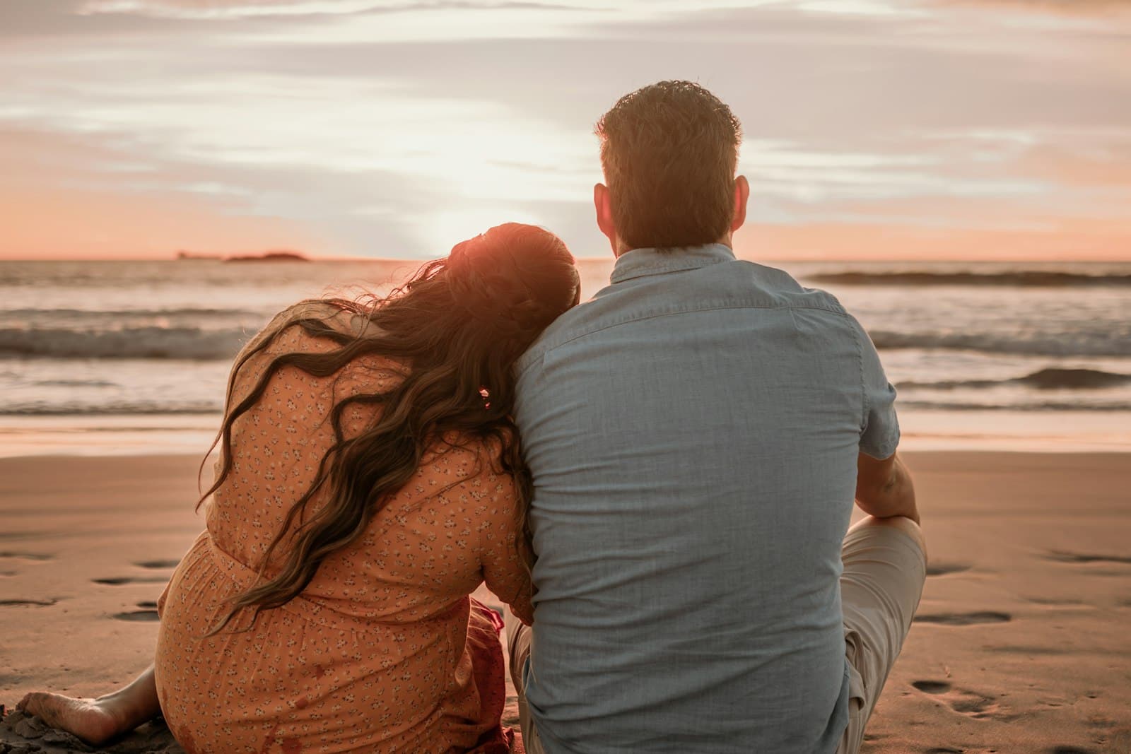 A couple sitting close on a beach at sunset, her head resting on his shoulder