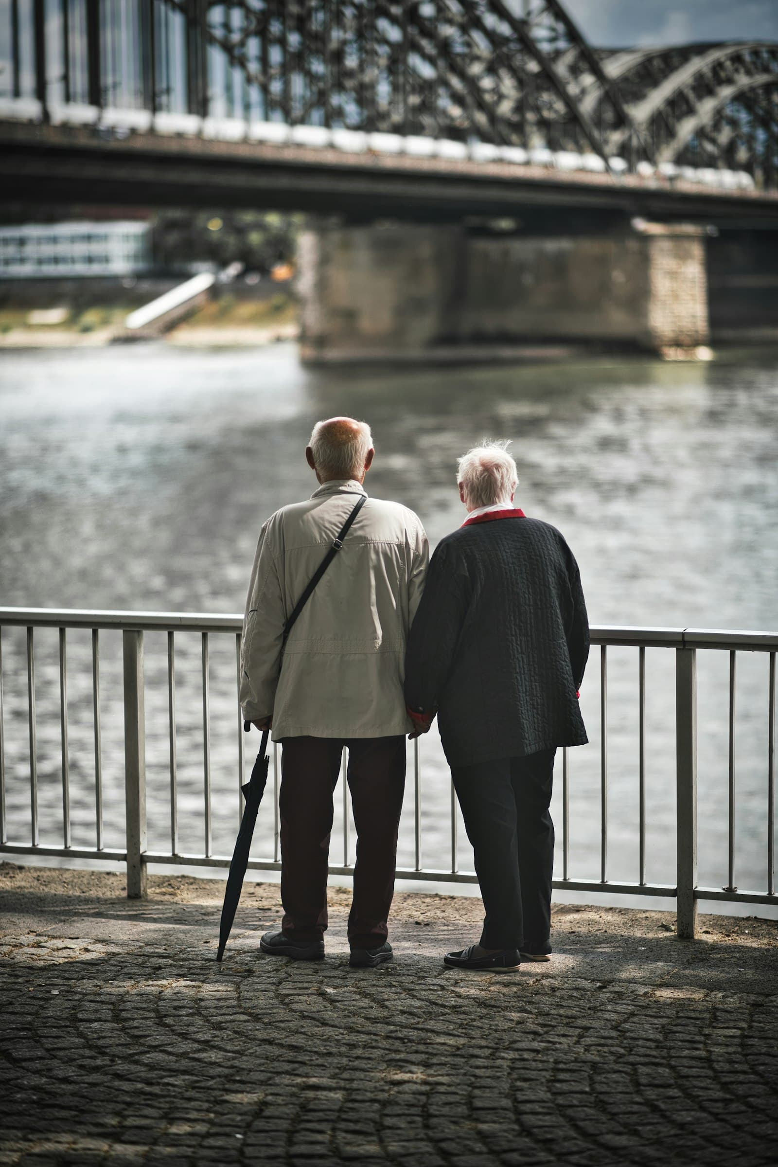An older couple standing side by side at a riverside railing, seen from behind