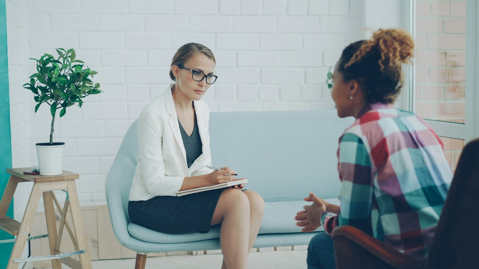 A therapist with a notepad listening to a client on a pale blue sofa in a bright office