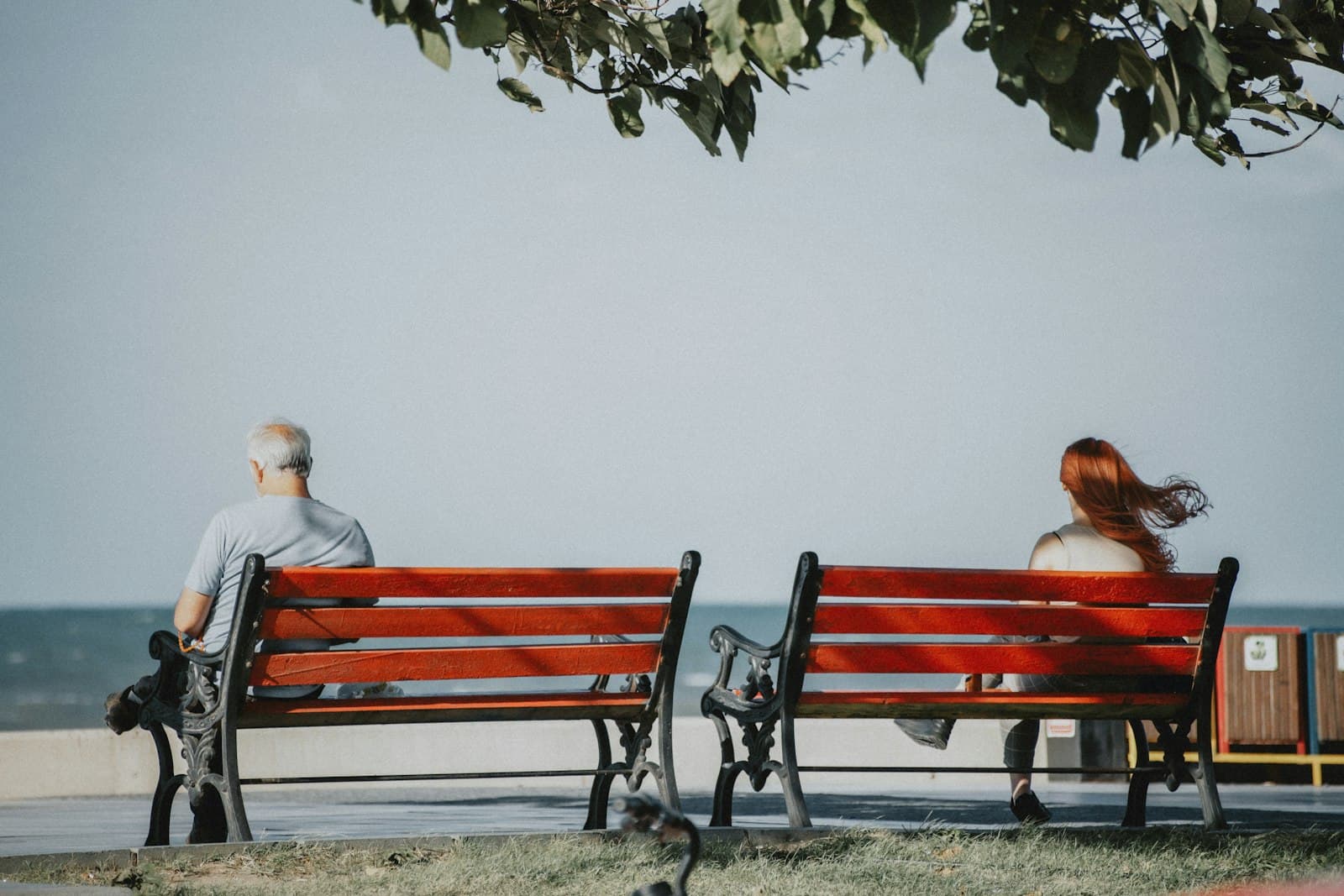 A man and a woman sitting on two separate red benches, facing the sea