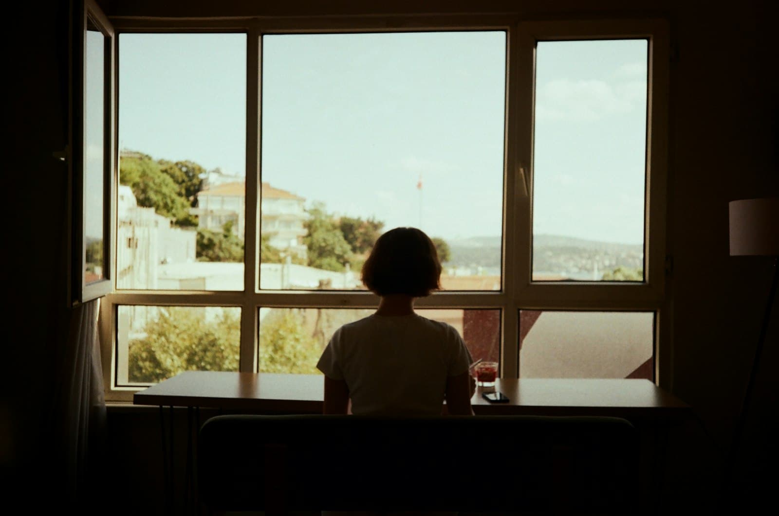 A woman sitting alone at a table by a large window in a dim room