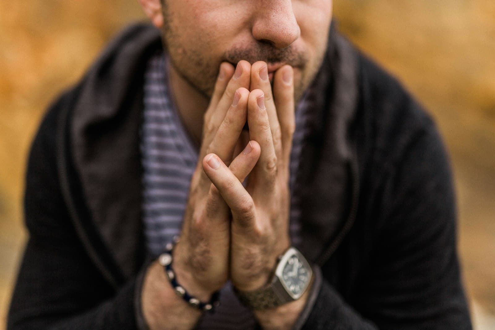 A bearded man with his hands pressed together against his mouth, looking down