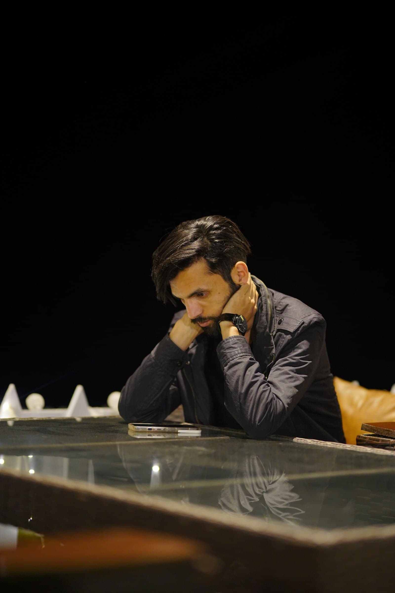 A man sitting alone at a glass table in a dark room, resting his head on his hand