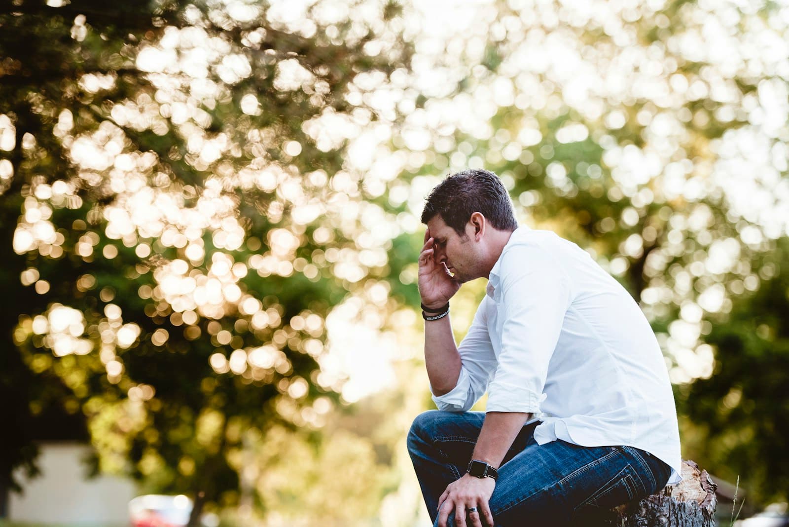 A man in a white shirt sitting outdoors under trees with his hand pressed to his forehead