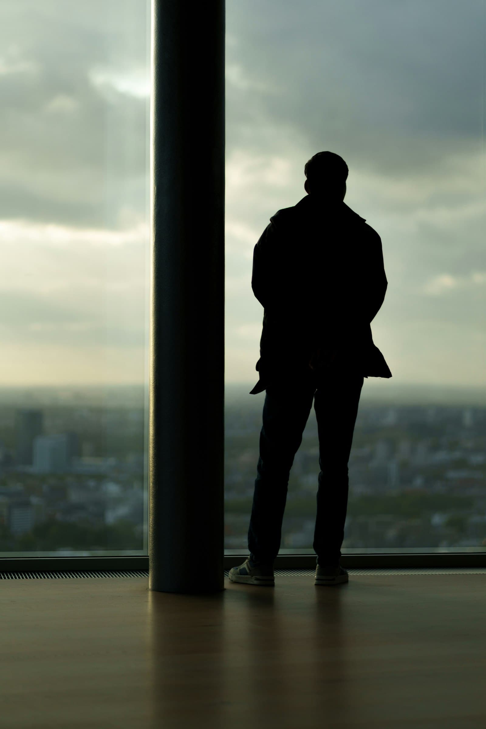 The silhouette of a man standing alone at a tall window overlooking the city