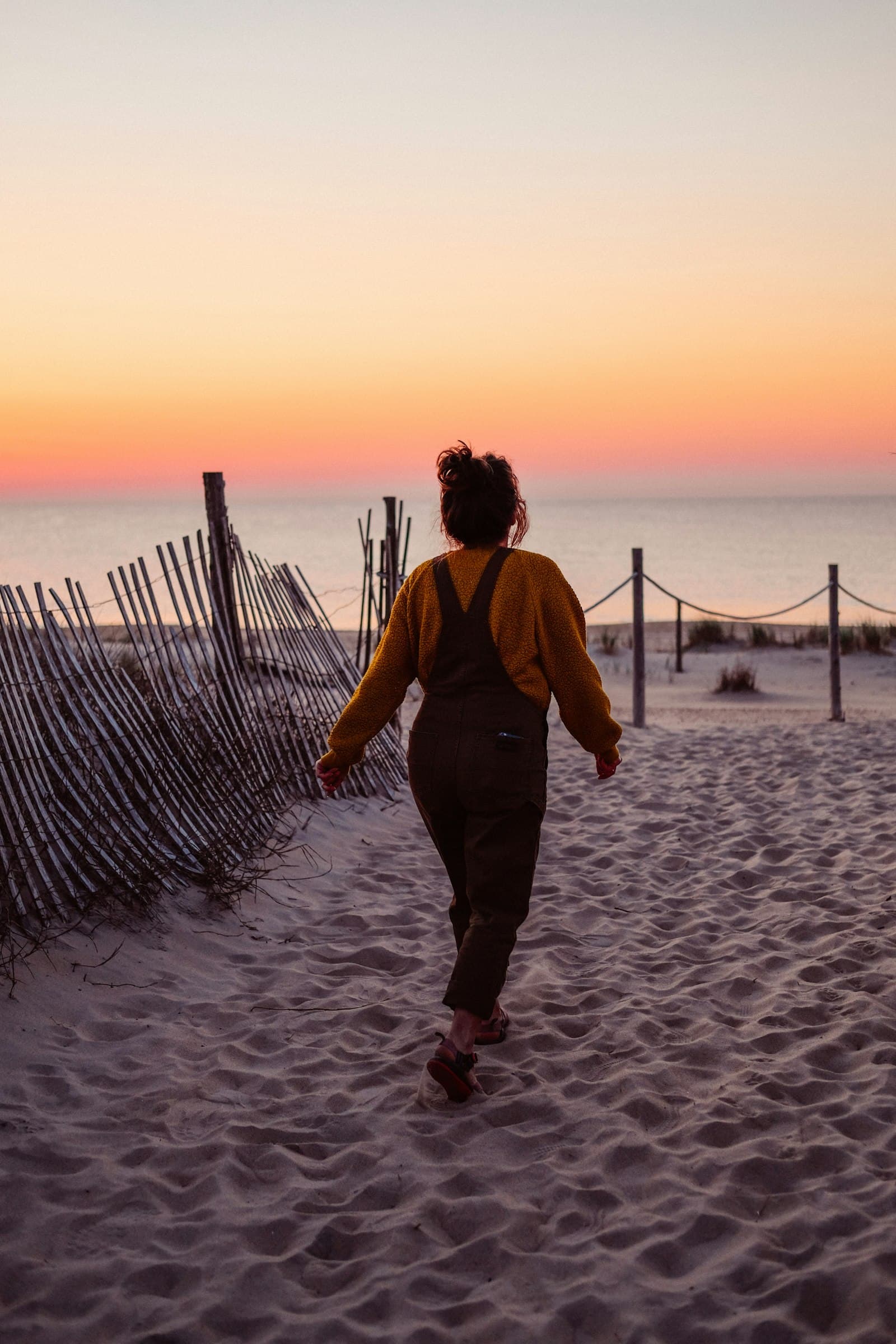 A woman walking alone along a sandy beach path at sunset, seen from behind