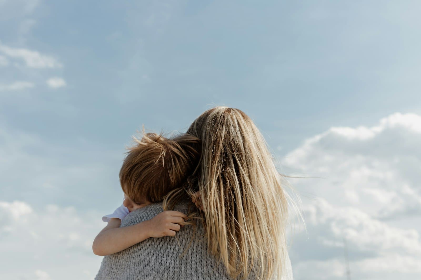 A child hugging a woman with long hair, seen from behind against a cloudy sky