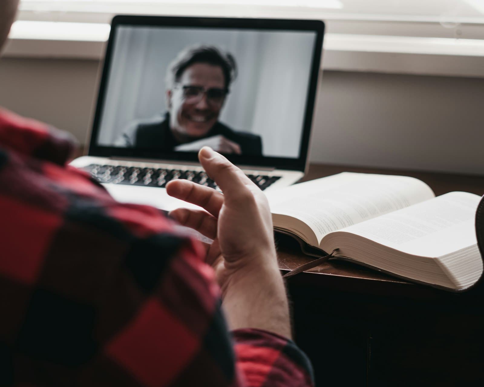 A person in a plaid shirt gesturing toward a smiling man on a laptop video call, an open book beside it