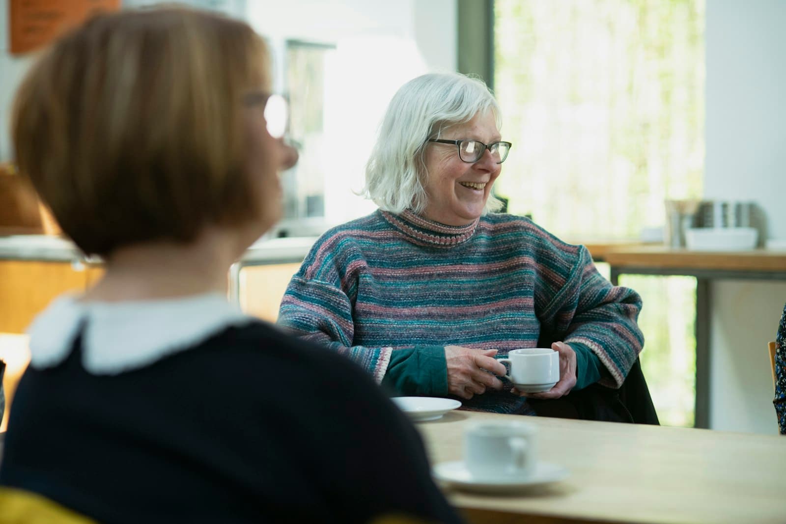 An older woman with white hair and glasses smiling over a cup of coffee as she talks with a friend
