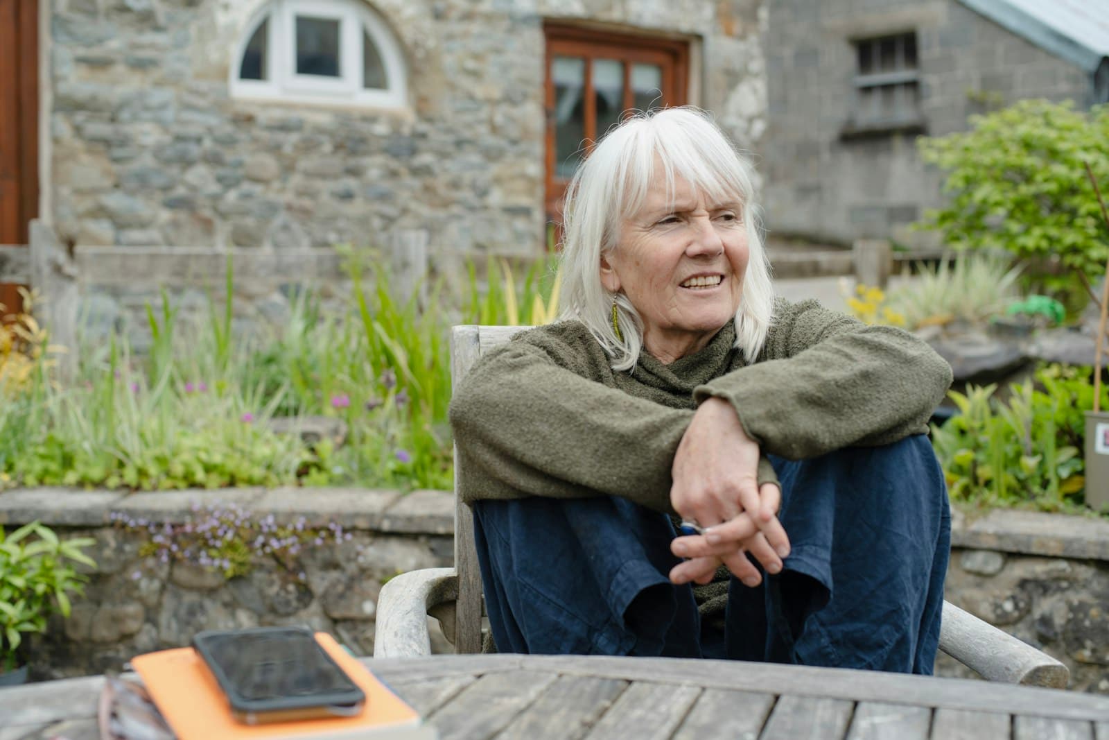 A smiling older woman with white hair sitting in a garden outside a stone house