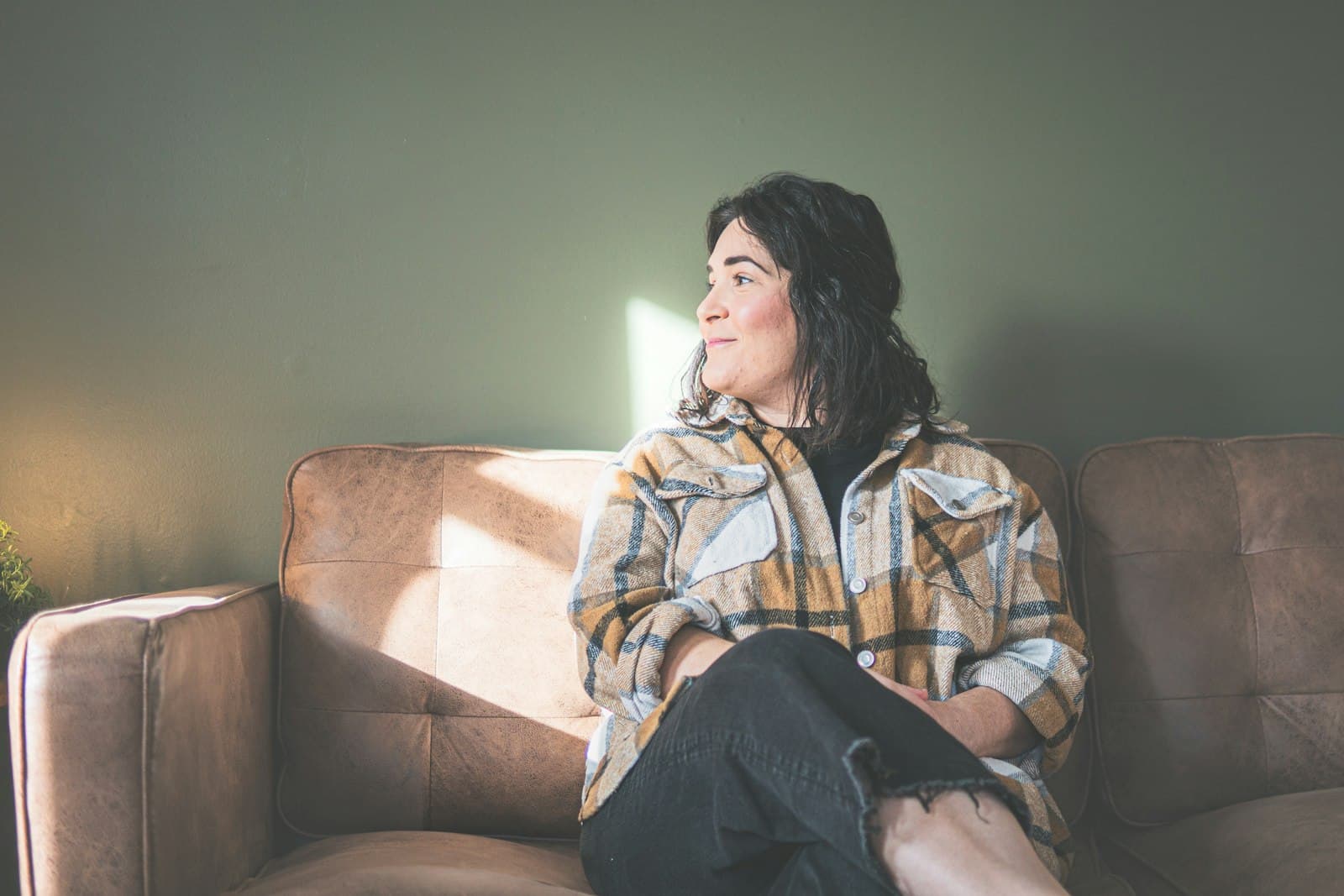A woman in a plaid shirt sitting on a leather sofa, gazing toward a sunlit window
