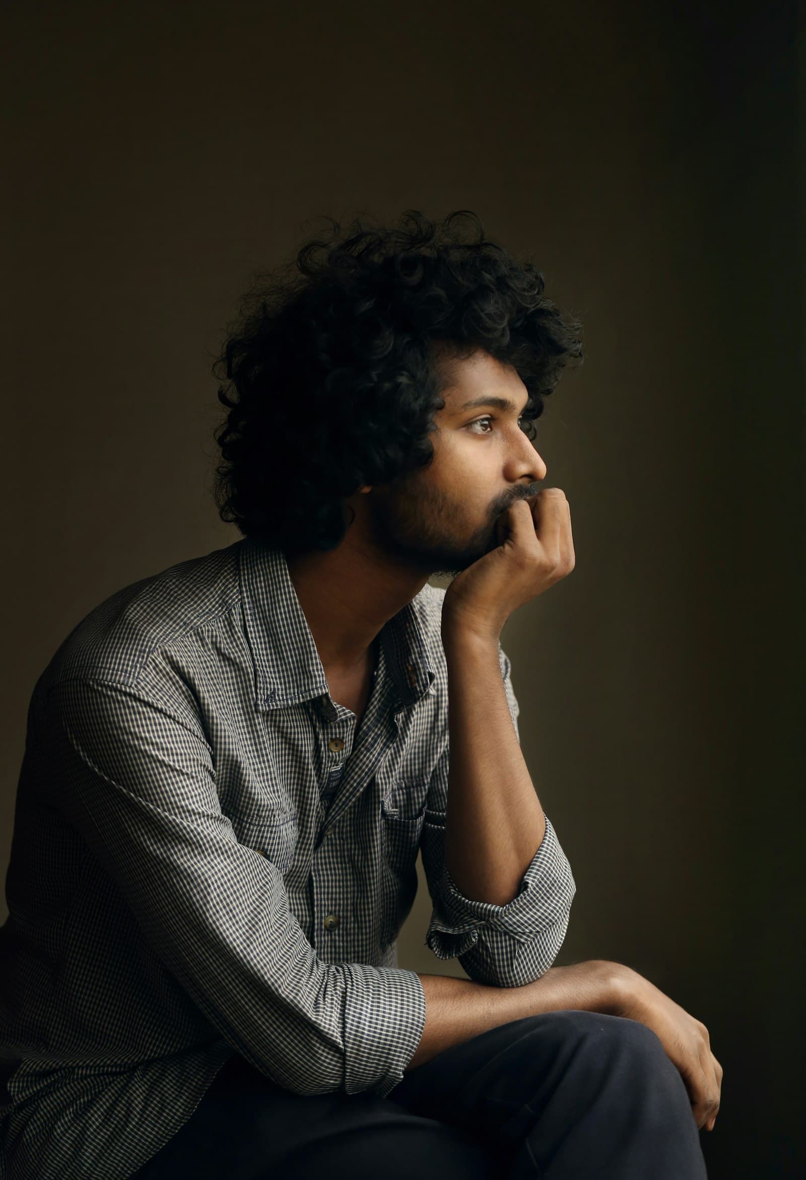 A young man with curly hair sitting in a dim room, hand at his mouth, deep in thought