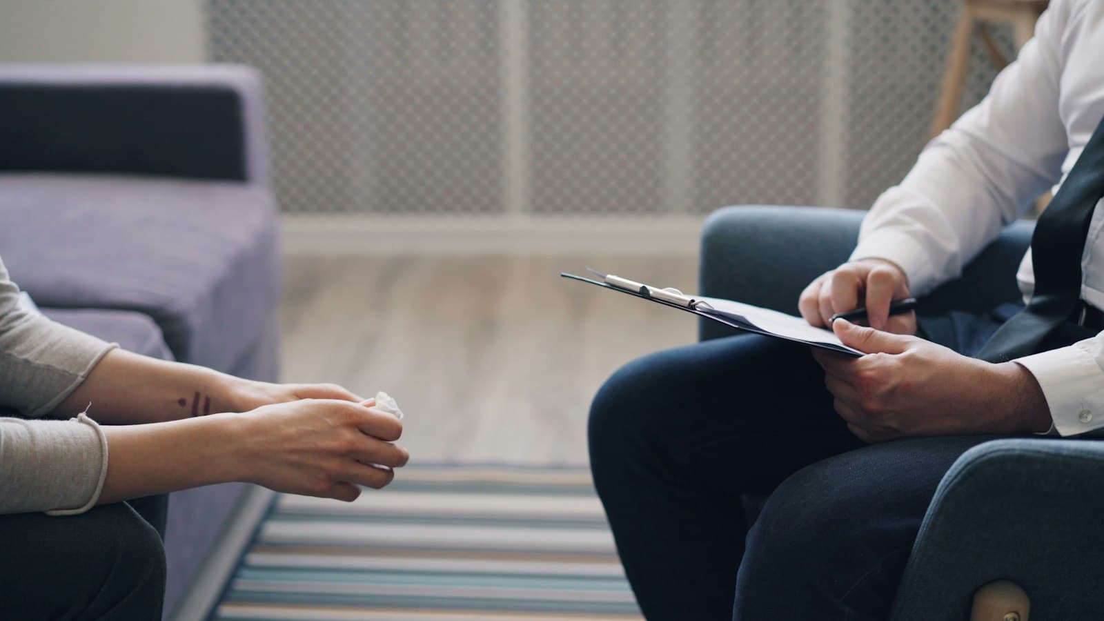 A therapist holding a clipboard and pen across from a client whose hands rest in her lap