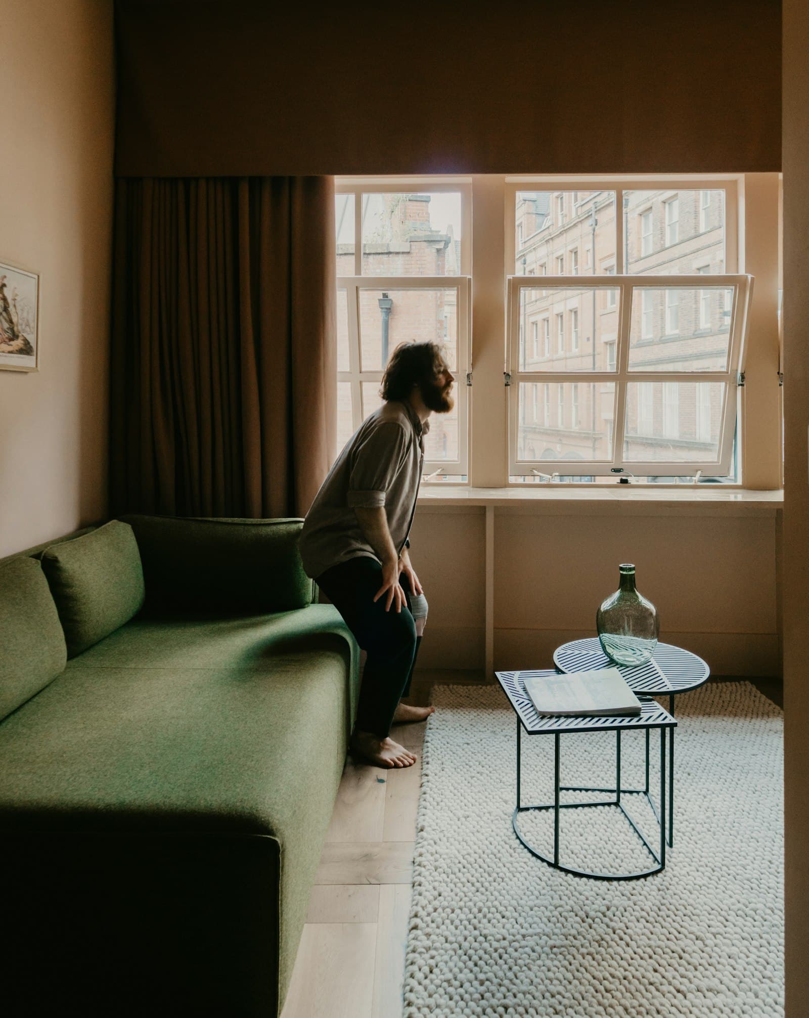 A man sitting alone on the arm of a green sofa, looking out a window in a quiet living room