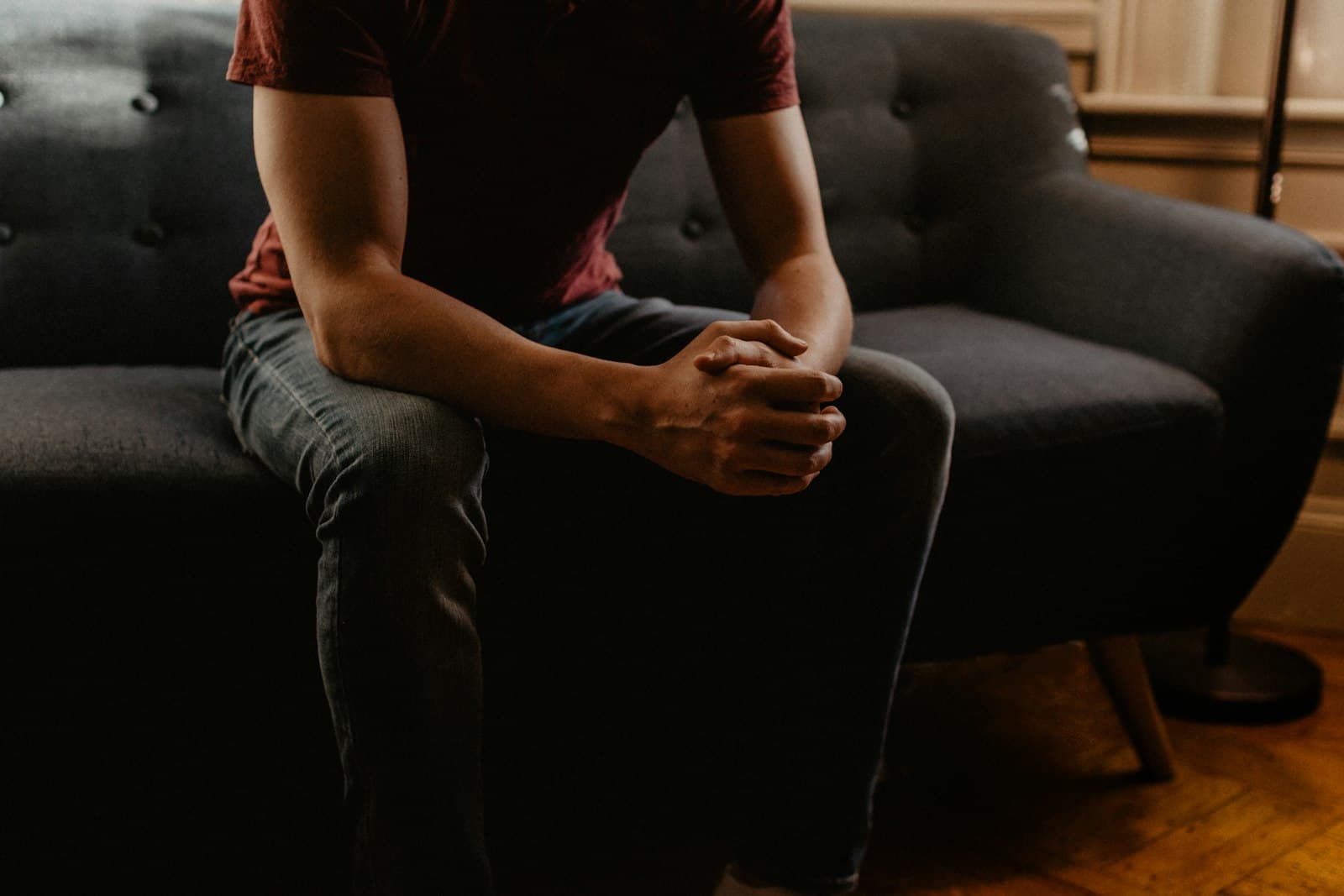 A man sitting on the edge of a dark sofa with his hands clasped between his knees