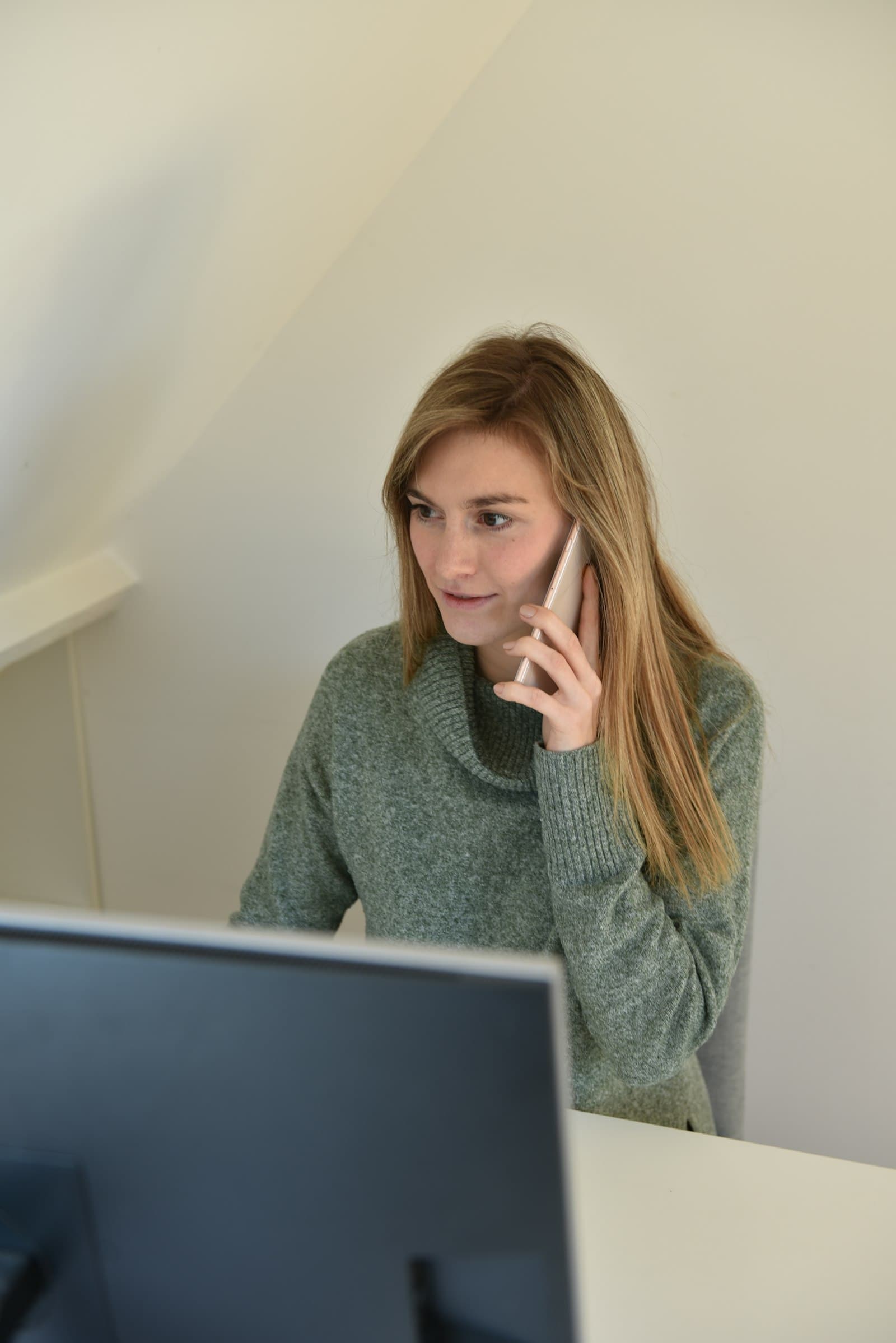A woman in a green sweater on a phone call at her desk, looking thoughtful