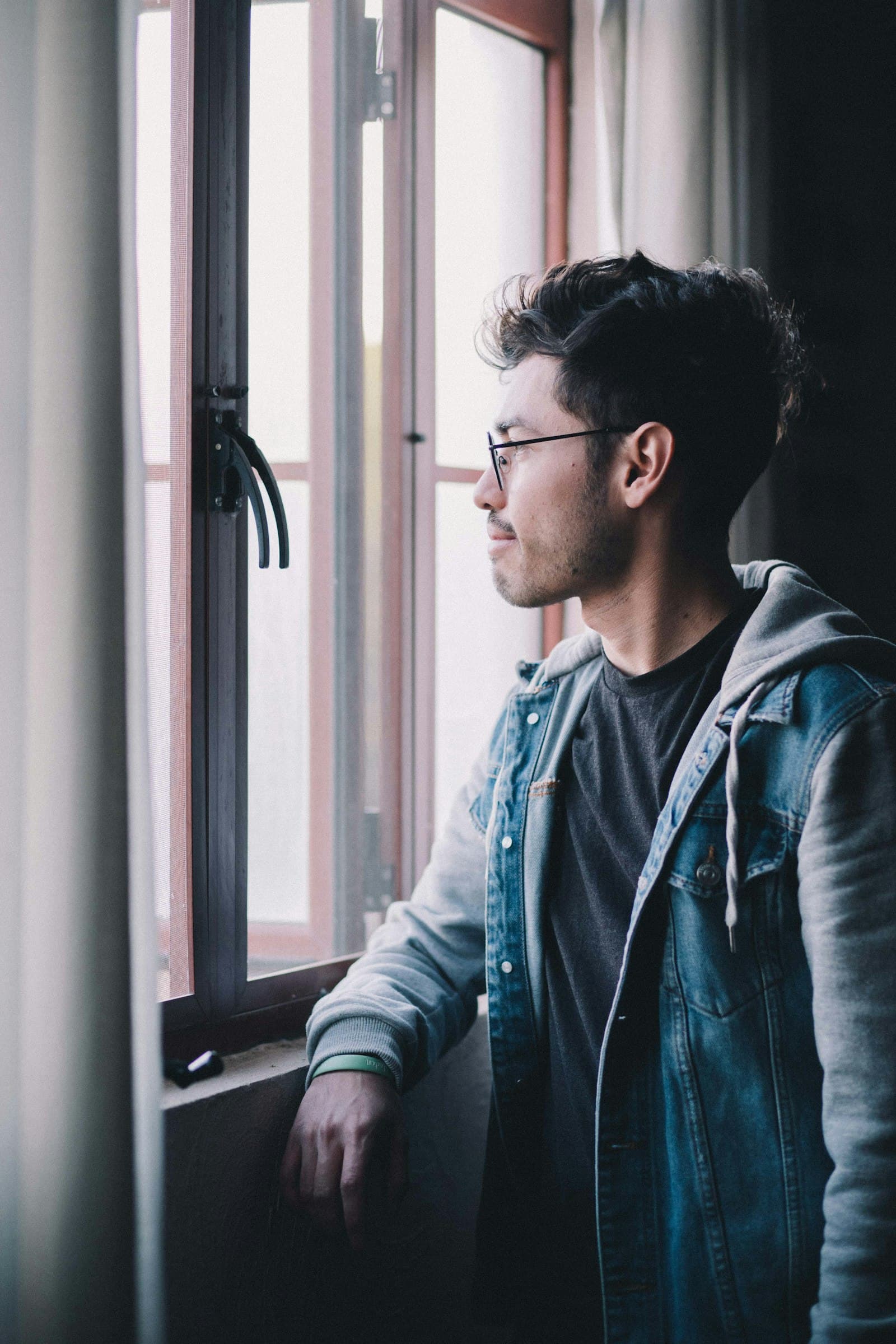 A young man in a denim jacket standing by a window, looking outside
