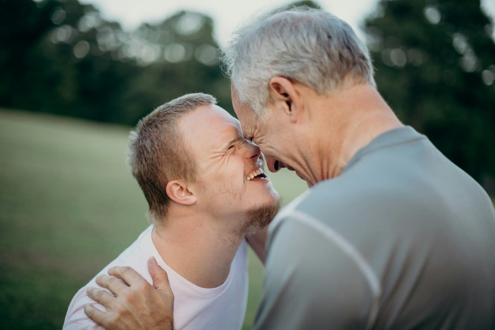 An adult son and his gray-haired father touching foreheads and laughing outdoors