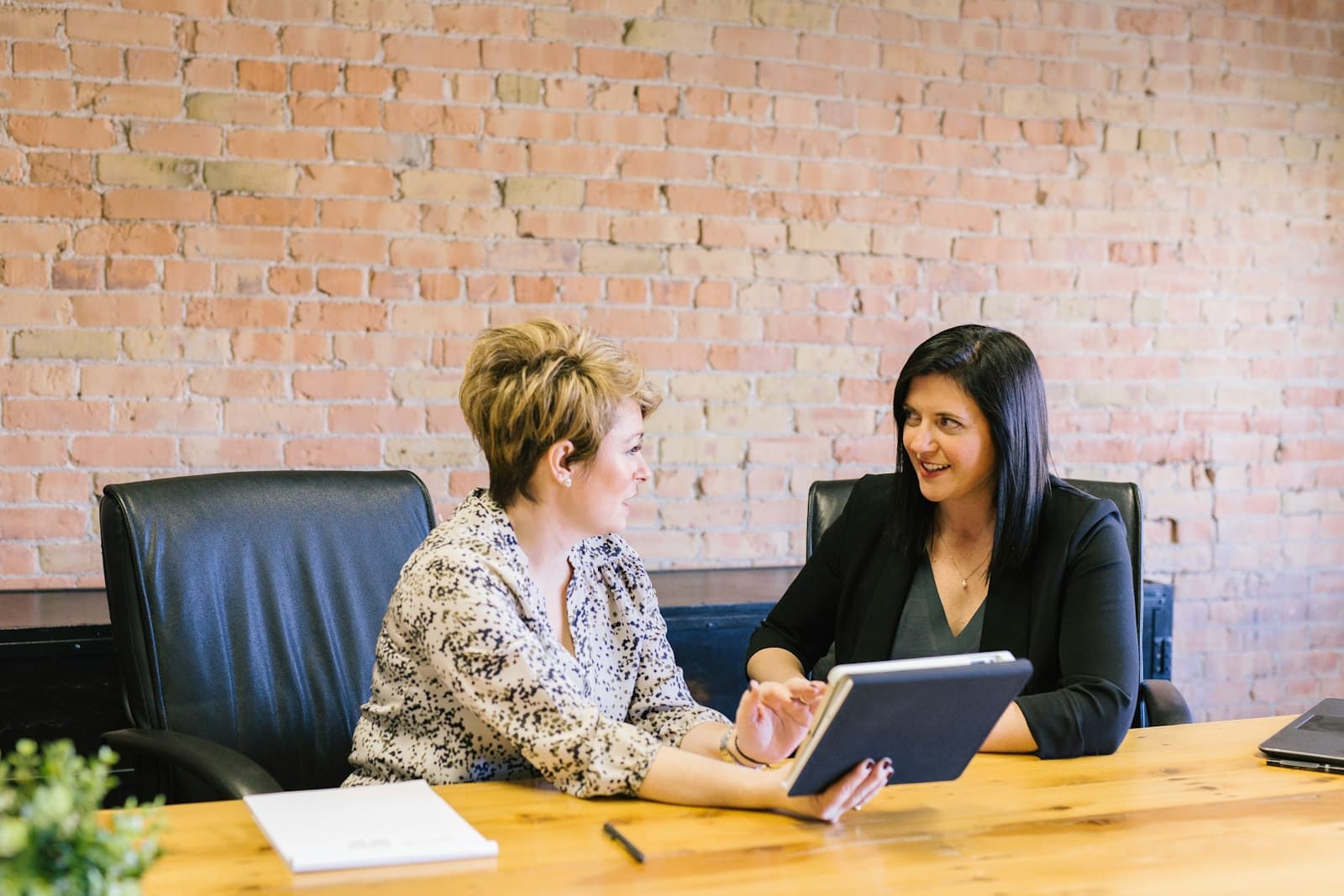 Two women talking at a wooden table, one holding a tablet
