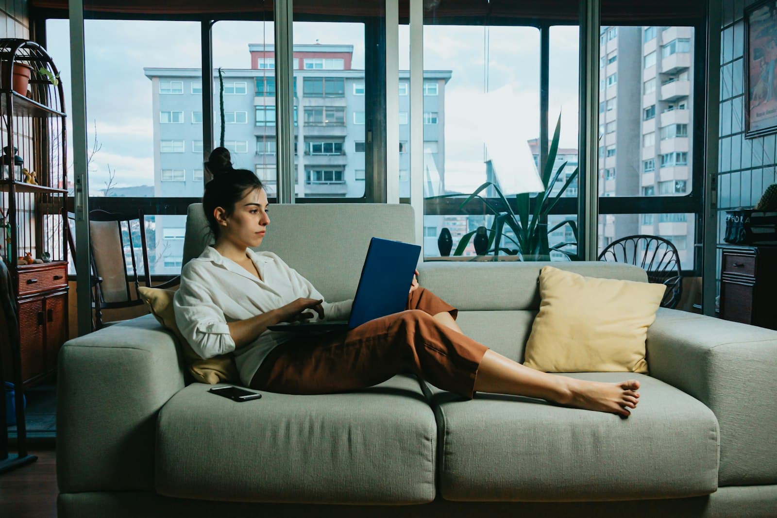 A woman working on a laptop on her sofa by large apartment windows