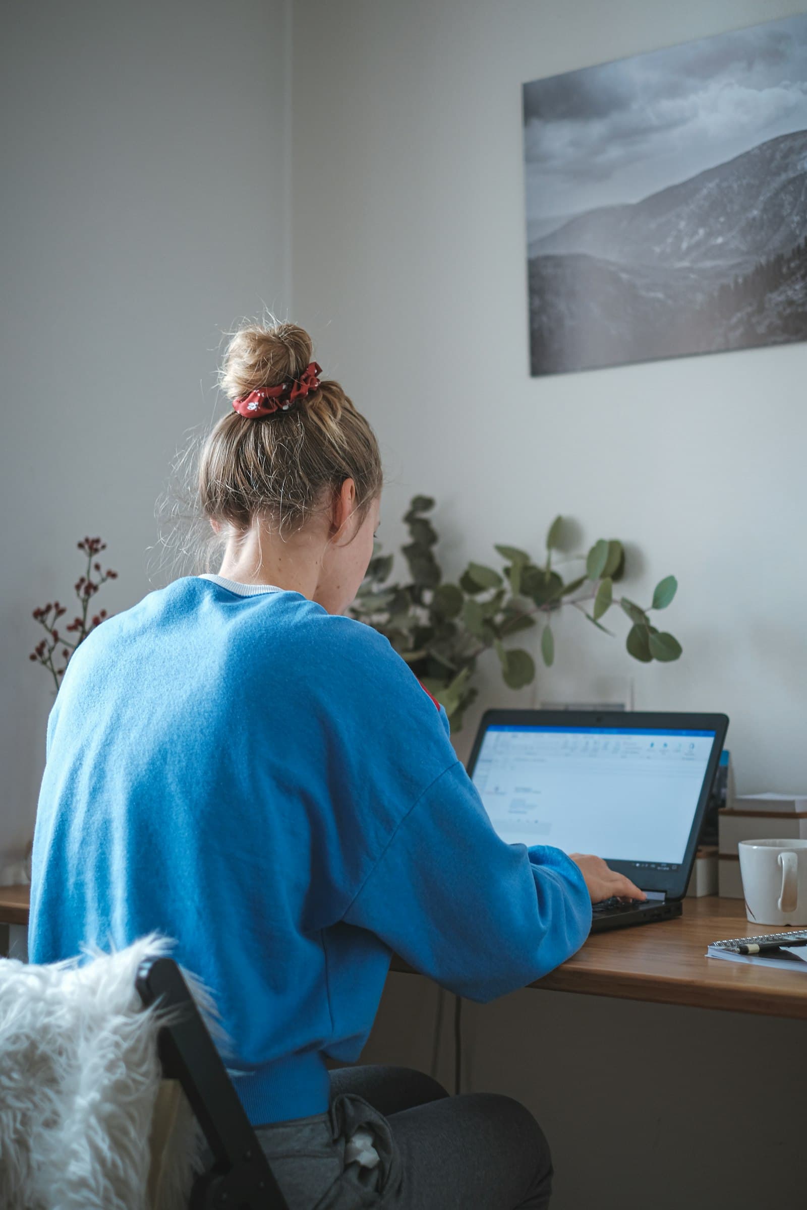 A woman in a blue sweater working at a laptop at a home desk, seen from behind